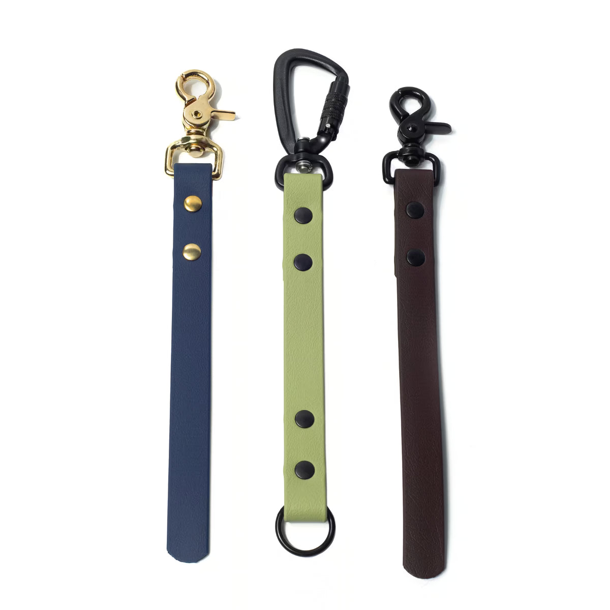 Biothane training tabs are shown in matcha with a matte black carabiner, espresso with a matte black trigger snap, and a deep sea blue with a solid brass trigger snap on a white background