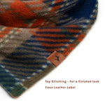 A close-up of a soft cotton fleece dog bandana with a plaid pattern in rust orange, navy blue, gray, and green, shown on a white background with a small faux leather label and text reading 'Top Stitching - For a finished look' and 'Faux Leather Label'.